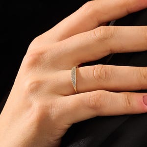 May include: A thin gold ring featuring a slightly tapered, raised center section that is pavé-set with numerous small, clear, sparkling stones. The band is smooth and polished, showcasing a delicate design that sits comfortably on the finger.