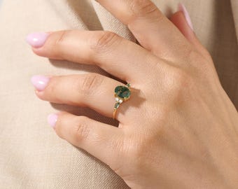 May include: A gold ring featuring an oval-shaped moss agate stone as the centrepiece, flanked by two small, clear, round-cut accent stones on each side. The band is thin and polished, showcasing a delicate design with a natural green gemstone.