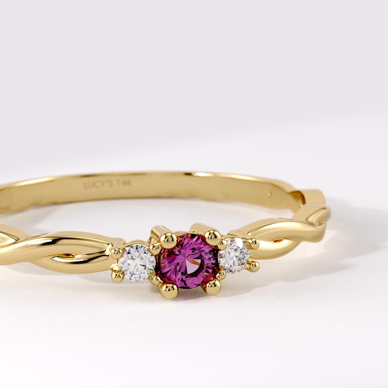 Pode incluir: Um anel de ouro amarelo de 14 quilates com uma pedra preciosa rosa de corte redondo central ladeada por duas pedras redondas transparentes menores. A banda tem um design torcido e est&aacute; gravada com o texto LUCY'S 14K na superf&iacute;cie interna.