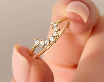 Puede incluir: Un anillo fino de oro con una banda curva en forma de V engastada con cinco piedras pequeñas, redondas y transparentes. La superficie interior de la banda tiene grabado el texto 14K, indicando la pureza del metal de esta joya.