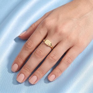 May include: A gold signet ring featuring a smooth, polished surface with a single small, round, embedded red gemstone. The ring has a tapered band and is worn on the ring finger, showcasing a minimalist design with a clean, reflective finish.