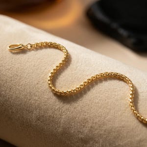 May include: A gold-colored chain necklace with a box link design. The necklace is displayed on a beige surface, with a lobster clasp closure. The chain is made of small, square links that create a textured appearance.