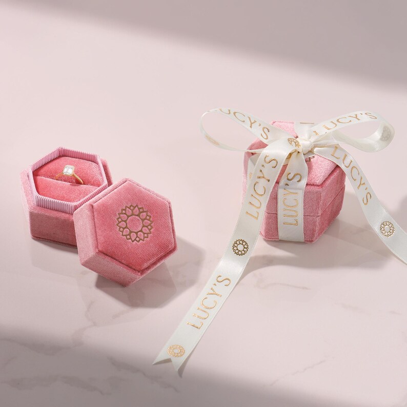 K&ouml;nnte beinhalten: Eine rosafarbene Samt-Ringbox mit sechseckiger Form und einem goldenen Blumenmuster. Die Box ist ge&ouml;ffnet und ein Ring befindet sich darin. Die Box steht auf einer wei&szlig;en Oberfl&auml;che. Ein wei&szlig;es Band mit dem Text "Lucy's" ist um eine kleinere rosafarbene Samtbox gebunden.