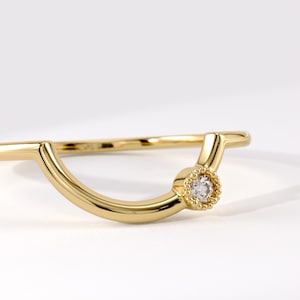 May include: A gold ring featuring a curved, U-shaped band with a single round, clear gemstone set in a beaded, circular bezel. The polished metal band has a smooth finish, and the setting is positioned asymmetrically along the curved arc.