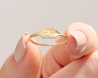 Può includere: Un anello in oro con una superficie rettangolare piatta caratterizzata da un motivo a raggiera di linee incise che partono dal centro. L'interno della sottile fascia presenta l'incisione LUCY'S 14K, che indica la purezza del metallo del gioiello.