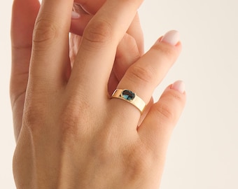 May include: A gold band ring featuring a single oval-shaped blue gemstone set in a bezel setting. The ring has a smooth, polished finish and a flat profile, showcasing a deep blue stone centered on the band, providing a clean and simple aesthetic.