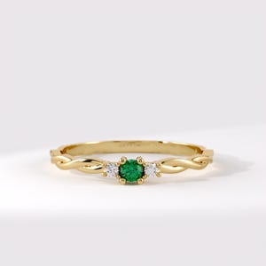 Peut inclure: Une bague en or avec une pierre précieuse verte ronde au centre, entourée de deux petites pierres claires rondes. L'anneau présente un motif torsadé en forme de corde et est fabriqué en or jaune poli, offrant une apparence délicate et raffinée.