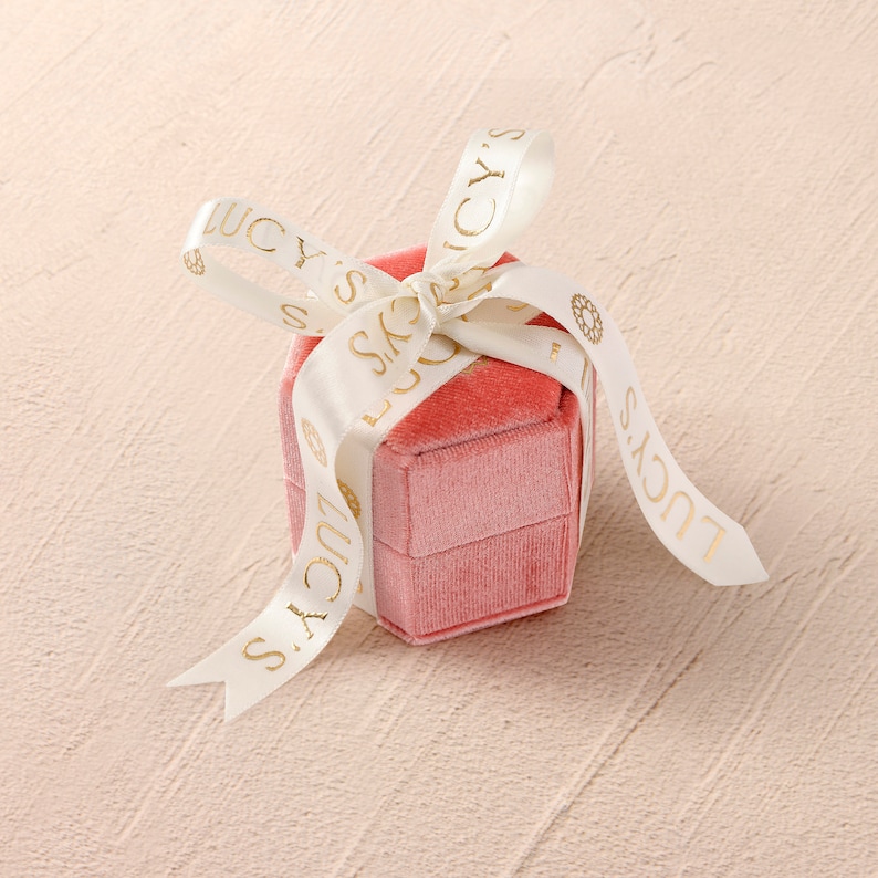 the ring box that products will be sent in is in a peachy pink color with creamy colored ribbon tied as a bow.