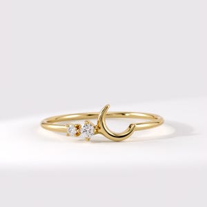 May include: A gold ring featuring a thin band with a crescent moon shape on one side and two small, round, clear stones set next to the moon. The metal has a polished, reflective finish, and the stones are positioned to catch the light.