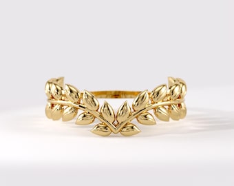 Könnte beinhalten: Ein goldener Ring mit einem V-förmigen Banddesign, das aus zwei symmetrischen Zweigen mit polierten Blättern besteht. Das Metall hat eine helle, reflektierende Oberfläche und die Struktur des Rings wölbt sich nach oben zu einem zentralen Punkt.