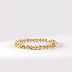 Op de afbeelding: Een gouden ring met een doorlopende band van kleine, gepolijste bolvormige kralen in een herhalend patroon. Het metaal heeft een heldere, reflecterende afwerking, wat zorgt voor een gestructureerd, cirkelvormig ontwerp met een uniforme vorm.