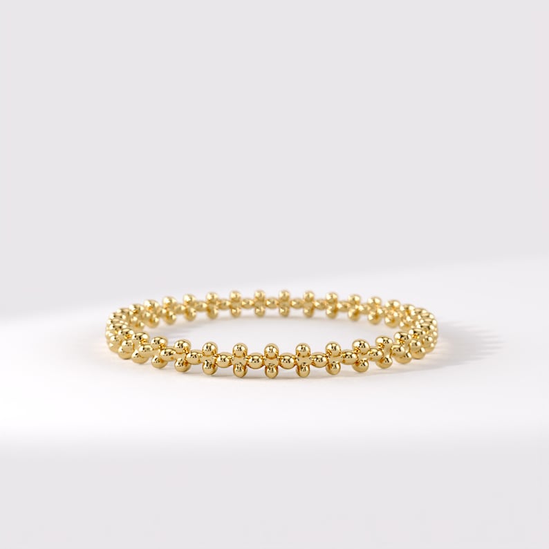 Op de afbeelding: Een gouden ring met een doorlopende band van kleine, gepolijste bolvormige kralen in een herhalend patroon. Het metaal heeft een heldere, reflecterende afwerking, wat zorgt voor een gestructureerd, cirkelvormig ontwerp met een uniforme vorm.