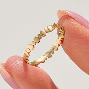 Könnte beinhalten: Ein goldener Ring mit einem sich wiederholenden Kieselsteinmuster. Der Ring wird zwischen zwei Fingern gehalten. Der Ring ist mit "LUCY'S 14K" gestempelt. Ein zarter Schmuck.