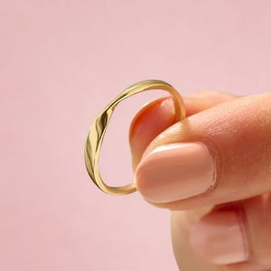 May include: A thin, polished gold ring with a smooth, continuous, and slightly irregular wave-like band. The metal surface is reflective and captures light, creating subtle highlights along the curved edges of the circular jewellery piece.