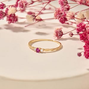 Puede incluir: Un anillo de oro con una banda texturizada, con una gema roja central y dos gemas blancas más pequeñas. Delicadas flores rosas en el fondo. Una joya elegante.