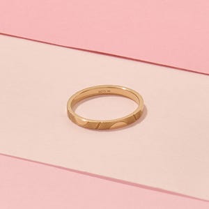 May include: A gold ring with a repeating sunburst pattern. The ring is thin and has the text "LUCY'S 14K" engraved on the inside. The ring is set against a pink background.