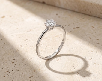 Op de afbeelding: Een zilverkleurige solitairring met een enkele, rondgeslepen heldere edelsteen in een zetting met zes pootjes. De dunne, gepolijste ringband is aan de binnenzijde gegraveerd met de tekst LUCY'S 14K en heeft een gladde, reflecterende afwerking.