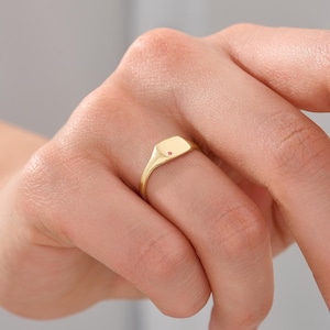 Könnte beinhalten: Ein goldener Siegelring mit einer glatten, flachen, rechteckigen Oberfläche, in deren Mitte ein einzelner, kleiner, runder, rosafarbener Edelstein eingelassen ist. Die Ringschiene ist schmal und poliert und weist durchgehend einen Goldton auf.