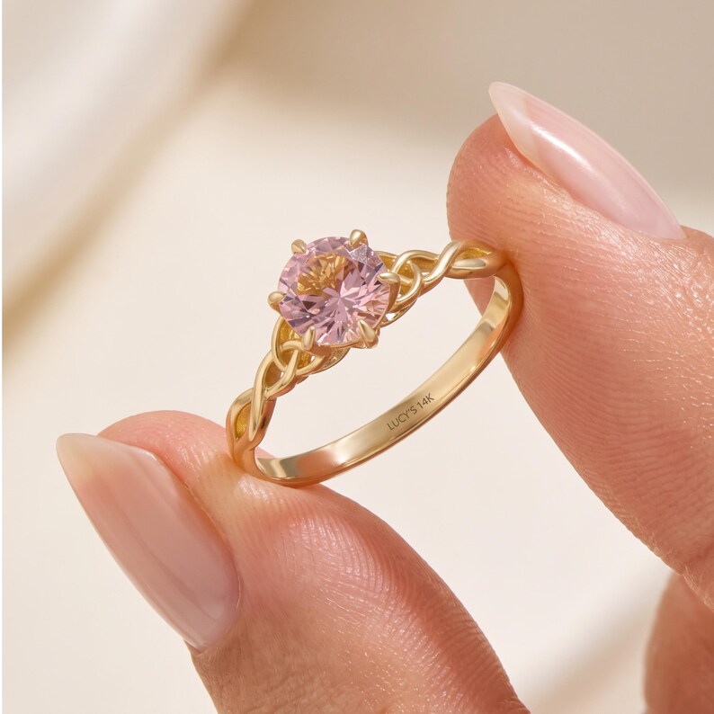 May include: A gold ring featuring a round, light pink gemstone set in a six-prong setting. The band has an intricate, twisted metal design on both sides of the stone. The interior of the band is clearly engraved with the text LUCY'S 14K.