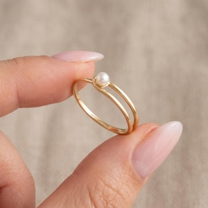 May include: A gold ring featuring a single small white pearl set at the top of a bypass-style band. The interior of the thin gold band is clearly engraved with the text LUCYS 14K, indicating the metal purity of the jewelry piece.