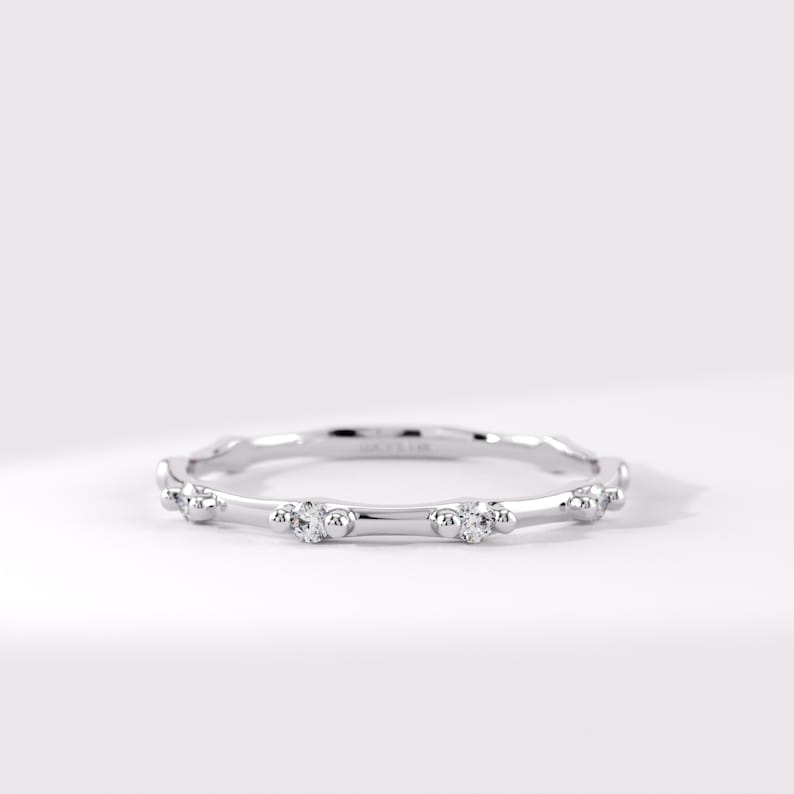 May include: A thin silver-toned metal band ring featuring a series of small, clear, round-cut stones set at intervals. Each stone is flanked by two tiny metal spheres, creating a repeating pattern along the circumference of the circular ring.