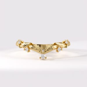 May include: A gold V-shaped ring featuring a series of textured leaf-shaped metal segments arranged in a chevron pattern, accented with five small, clear, round-cut stones set at intervals along the band's top edge.
