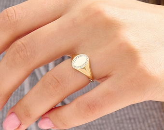 May include: A gold signet ring featuring an oval-shaped face with a polished center and a border of small, clear, sparkling stones set into the metal. The band is smooth and tapers slightly toward the back, showcasing a classic, clean design.