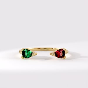 May include: A gold open ring featuring two pear-cut gemstones, one vibrant green and one deep red, set in polished gold prongs. The band is thin and circular, with the text LUCY 14K engraved on the inner surface of the gold metal band.