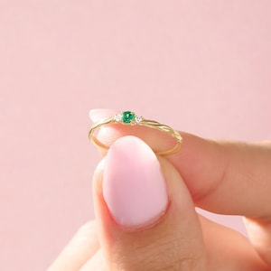 May include: A gold ring with a twisted band design, featuring a small, square-cut green gemstone in the center, flanked by two smaller white gemstones. The ring is held against a soft pink background.