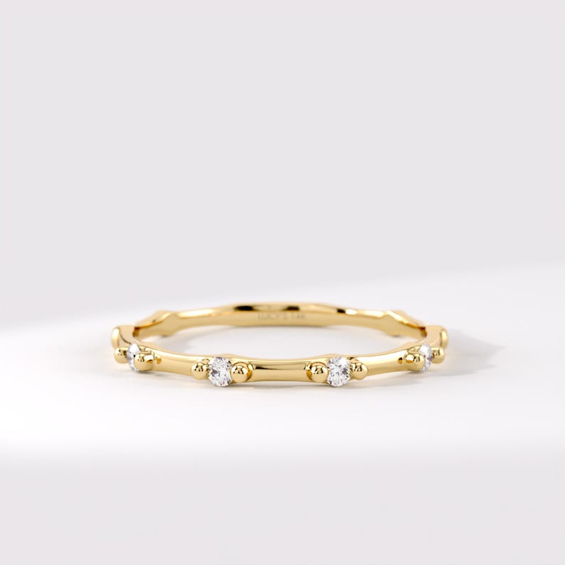 May include: A thin yellow gold band ring featuring a series of small, round, clear gemstones set at intervals. Each gemstone is flanked by two tiny gold spheres, creating a delicate, repeating pattern along the circumference of the polished metal ring.