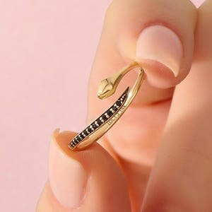 May include: A gold snake-shaped ring featuring a row of small black stones along the body and a smooth head. The inner band is engraved with the text LUCIS 14K. The design is open-ended, allowing for an adjustable fit around the finger.