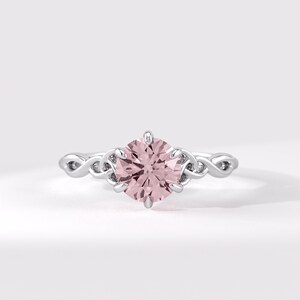 May include: A silver-toned ring featuring a round, pale pink center stone held by six prongs. The band is crafted with an intricate, continuous interwoven metal pattern that extends from the setting along both sides of the finger band.