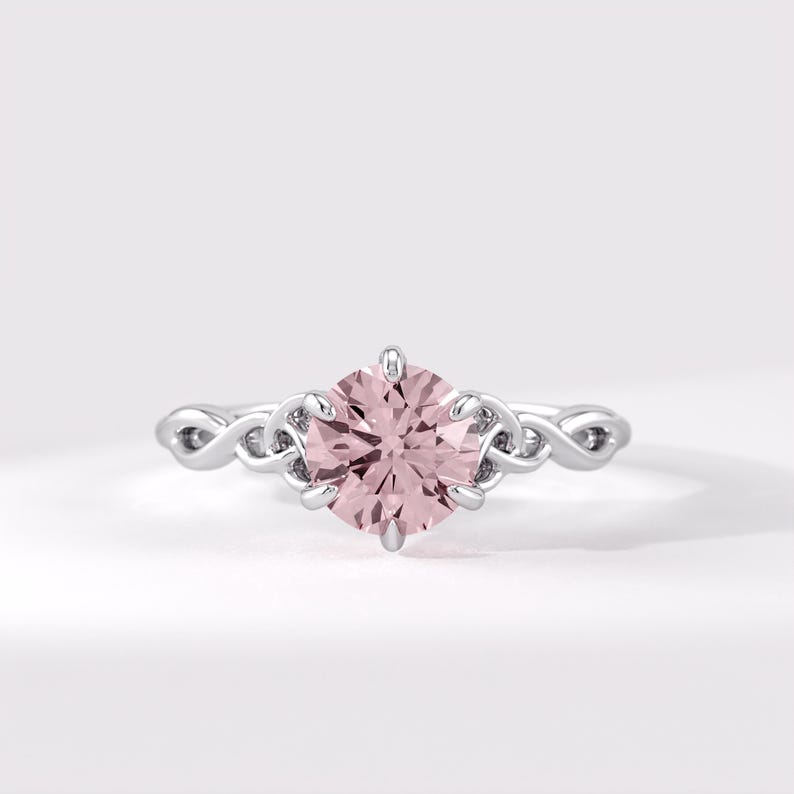 May include: A silver-toned ring featuring a round, pale pink center stone held by six prongs. The band is crafted with an intricate, continuous interwoven metal pattern that extends from the setting along both sides of the finger band.