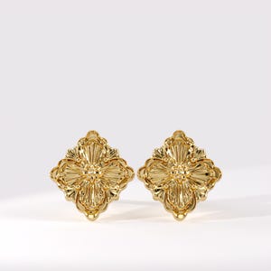 May include: A pair of gold-toned stud earrings featuring a symmetrical, diamond-shaped design with intricate, raised floral-inspired relief patterns and a polished, reflective metallic finish that catches the light across the textured surface.