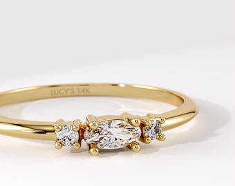 Op de afbeelding: Een gouden ring met een centrale marquise-geslepen heldere edelsteen, geflankeerd door twee kleinere rondgeslepen heldere edelstenen. De binnenzijde van de dunne, gepolijste gouden ring is voorzien van de gravure LUCY'S 14K voor metaalzuiverheid.