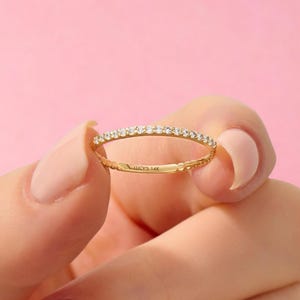 May include: A thin gold band ring featuring a row of small, clear, sparkling stones set along the top half. The interior of the band is clearly engraved with the text LUCY'S 14K, indicating the metal purity of the jewelry piece.