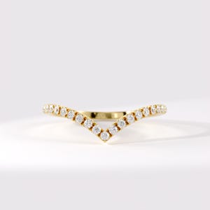 May include: A V-shaped gold ring featuring a row of small, round, clear, faceted stones set along the entire outer curve. The band has a polished metallic finish, and the stones are arranged in a continuous chevron pattern across the front.