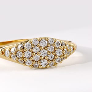 May include: A gold ring featuring a wide, oval-shaped face densely set with numerous small, round, clear, faceted stones. The stones are arranged in a cluster pattern, held by small gold prongs, creating a textured, sparkling surface on the band.