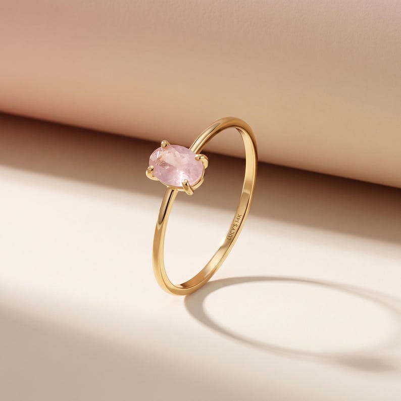 May include: A gold ring featuring a pale pink oval gemstone. The ring has a delicate band, and the gemstone is held in place by four prongs. The ring is presented on a light pink and white surface.