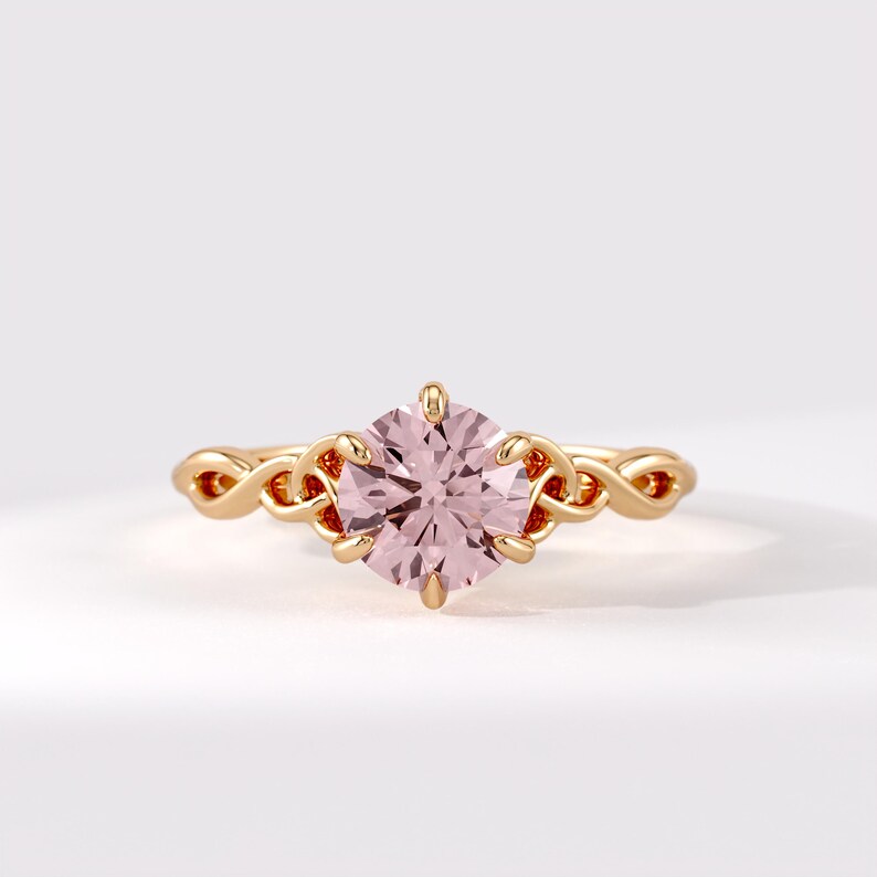 May include: A gold ring featuring a round, pale pink gemstone held by six prongs. The band is crafted with an intricate, interwoven knot design that extends from the central setting, showcasing a polished metallic finish and a delicate appearance.