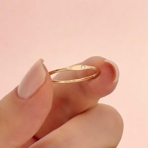 May include: A thin gold ring featuring a small, round, clear gemstone set into a flat, elongated oval section on the band. The interior of the band is engraved with the text LUCY'S 14K, indicating the metal purity of the gold ring.