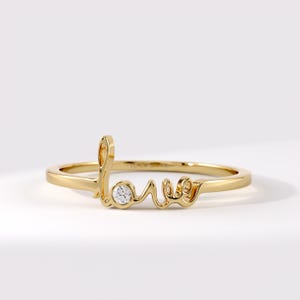 Könnte beinhalten: Ein goldener Ring mit dem Wort love in Schreibschrift. Der Buchstabe o ist durch einen kleinen, runden, klaren und facettierten Edelstein ersetzt, der in das Metallband eingelassen ist. Der Ring hat eine polierte Oberfläche und ein schmales Band.
