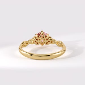 May include: A gold ring featuring a round, faceted pink gemstone set in a decorative, openwork gallery. The band is smooth and polished, transitioning into intricate, interlaced metalwork that supports the central stone setting.