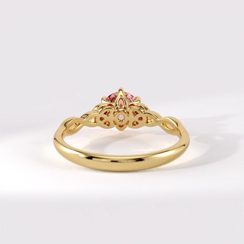 May include: A gold ring featuring a round, faceted pink gemstone set in a decorative, openwork gallery. The band is smooth and polished, transitioning into intricate, interlaced metalwork that supports the central stone setting.
