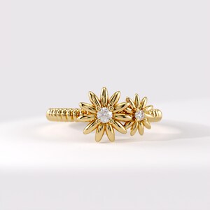 May include: A gold ring featuring a textured, ribbed band and two flower-shaped motifs of varying sizes. Each flower is crafted from smooth, rounded gold petals and centres on a single, clear, round-cut gemstone set in the middle of the bloom.