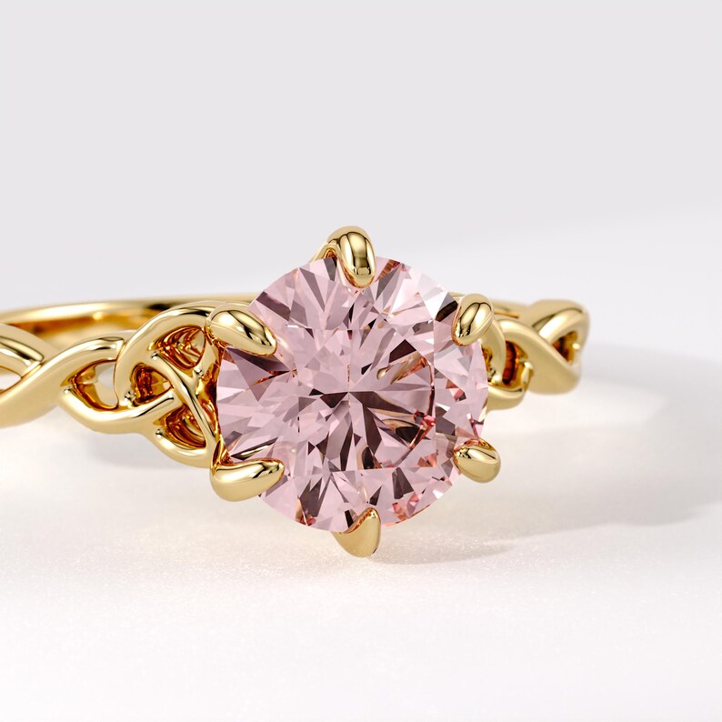 May include: A gold ring featuring a large, round, pale pink gemstone held by six prongs. The band is crafted with an intricate, interwoven Celtic-style knot pattern that connects to the central setting, showcasing a polished metallic finish.