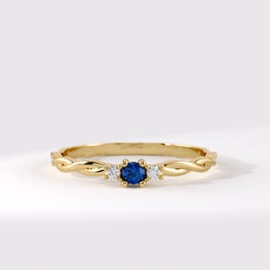 Op de afbeelding: Een geelgouden ring met een centrale ronde blauwe edelsteen, geflankeerd door twee kleinere ronde heldere stenen. De ringband heeft aan beide zijden van de stenen een gedraaid, touwachtig ontwerp. Het metaal is gepolijst en glanst in het licht.