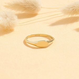 May include: A polished gold signet ring with a smooth, oval-shaped face. The inner band of the ring is clearly engraved with the text LUCY'S 14K, indicating the metal purity. The ring features a slim, rounded band that tapers toward the center.