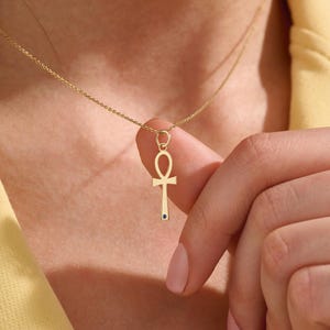 May include: A gold-colored ankh pendant necklace with a small blue gemstone at the bottom. The ankh symbol is suspended from a delicate chain. The necklace is worn around the neck.