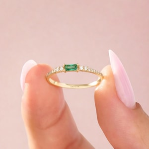 Puede incluir: Un delicado anillo de oro con una gema verde rectangular con corte esmeralda. La banda está adornada con pequeñas y brillantes gemas blancas. El anillo se sostiene sobre un fondo rosa suave. El texto "LUCY'S 14K" es visible en el anillo.